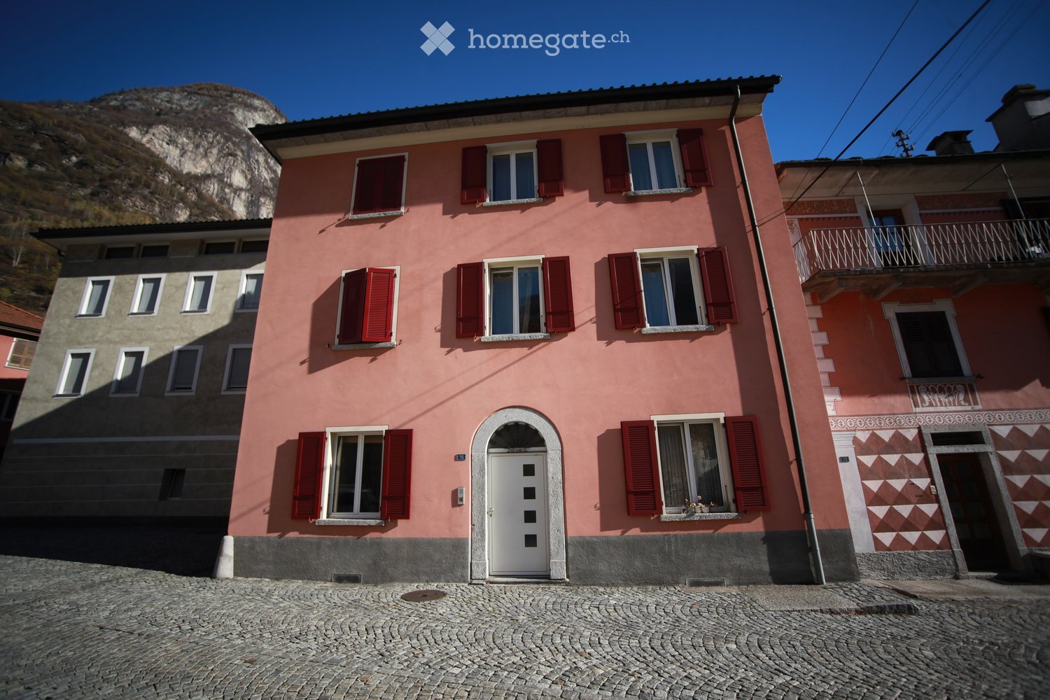 Two-story pink house with red shutters, white door, and balcony on the right side. The house is situated on a cobblestone street with a mountain in the background.