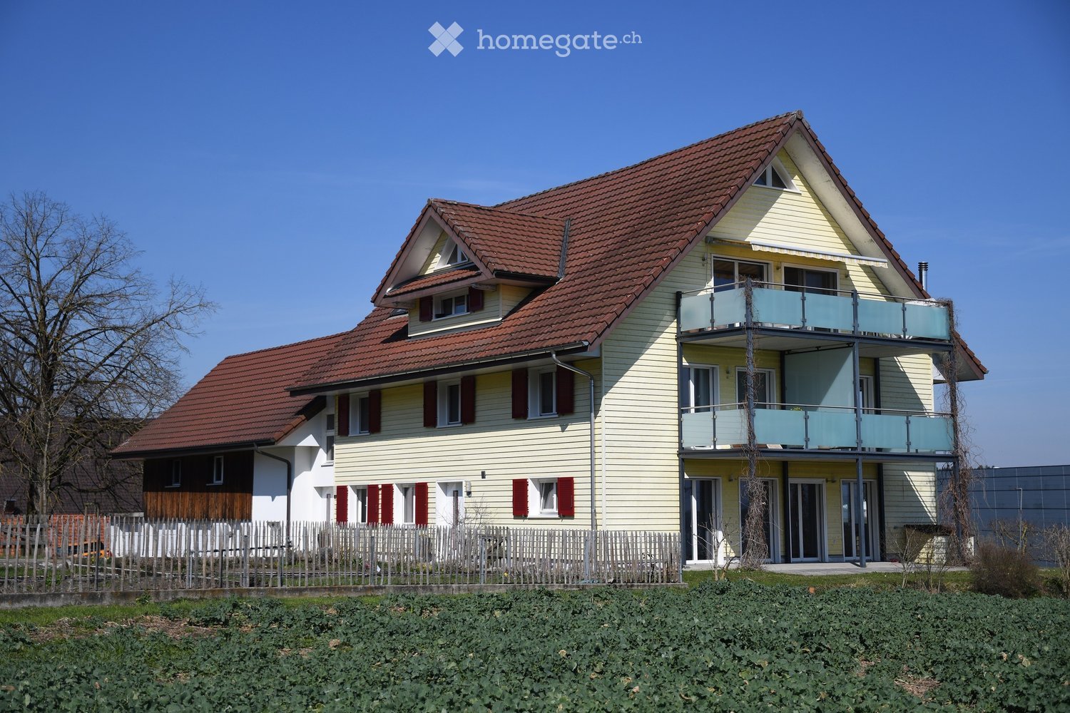 Two-story house with red-tiled roof, yellow siding, multiple windows, and a balcony on the upper level.