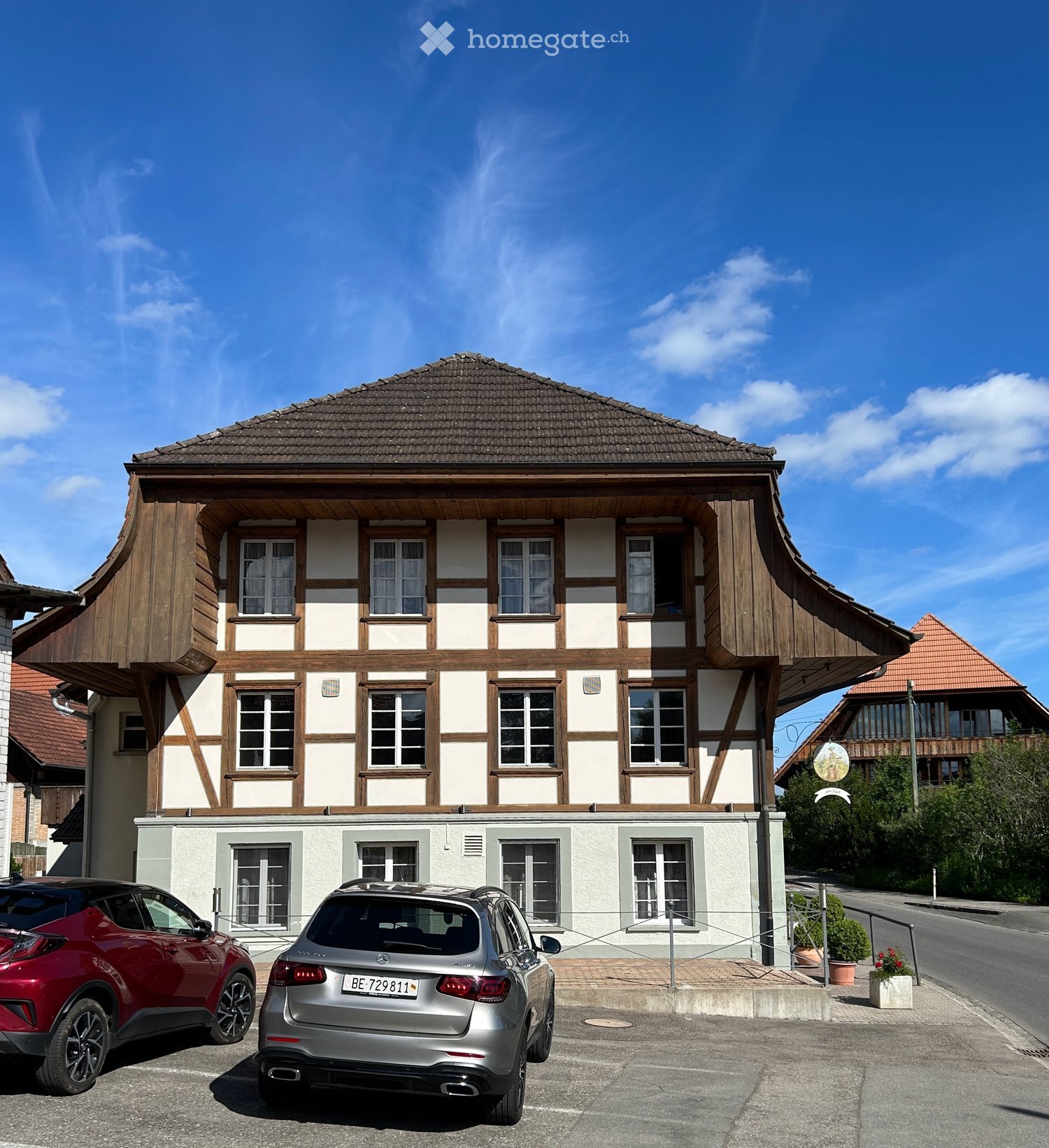 Two-story traditional half-timbered house with white plaster walls and wooden beams, multiple windows, and a tiled roof. Two cars are parked in front.