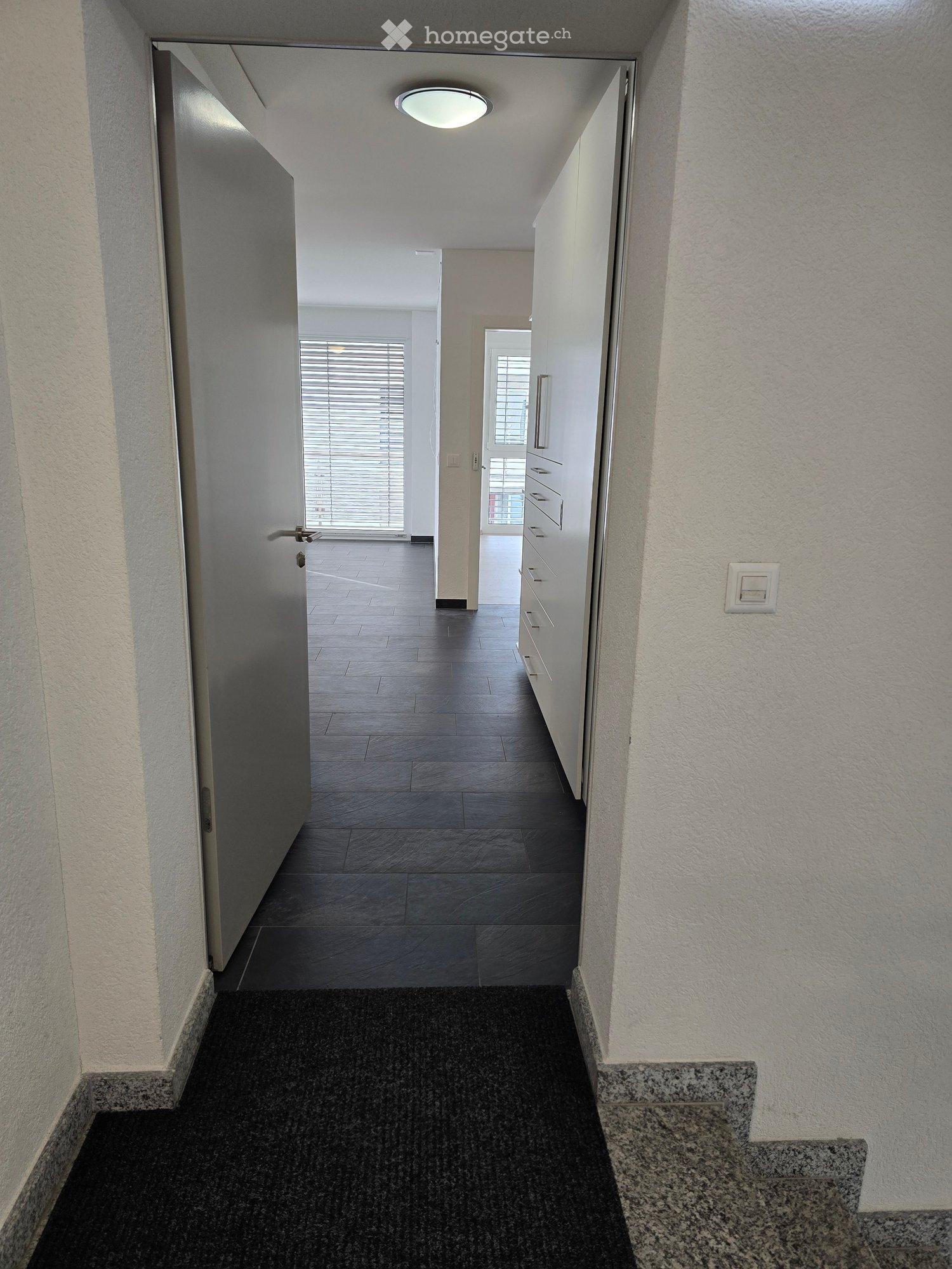 Clean, modern hallway with dark tiled floor, white walls, and a ceiling light. Features a partially open door leading to another room and a staircase with granite steps.