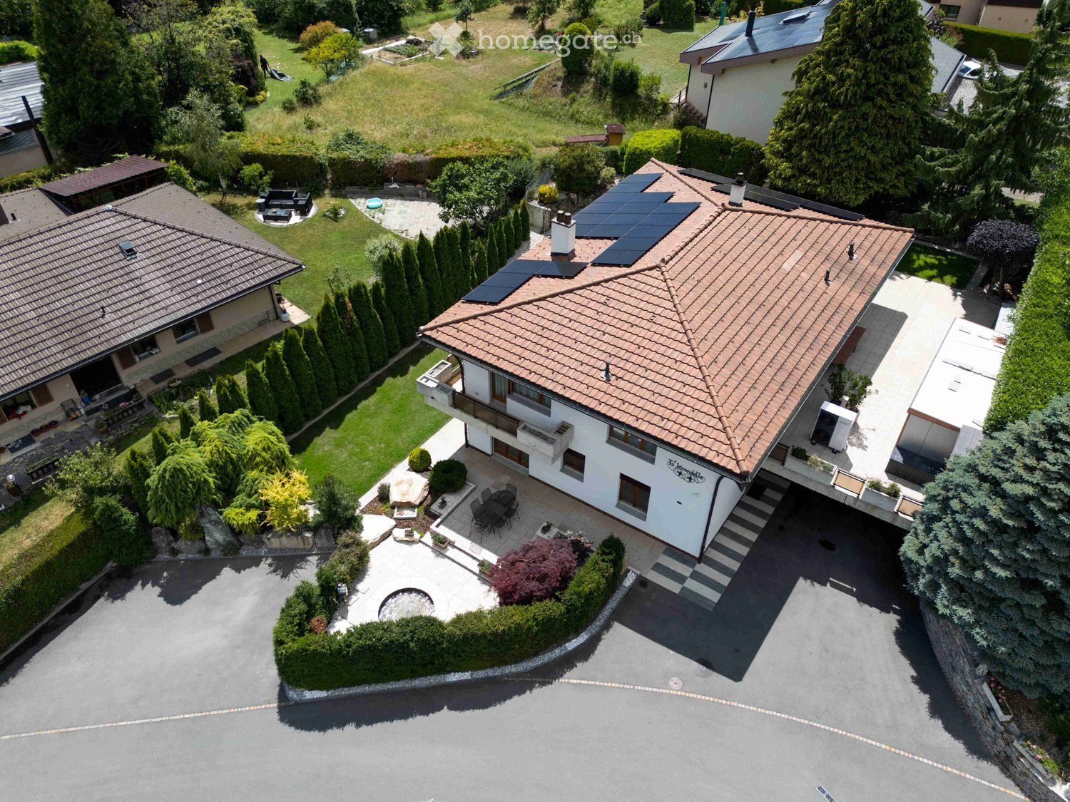 Aerial view of a two-story house with a tiled roof, surrounded by a well-maintained garden, patio, and outdoor seating area. The house features solar panels on the roof and a driveway leading to a garage.
