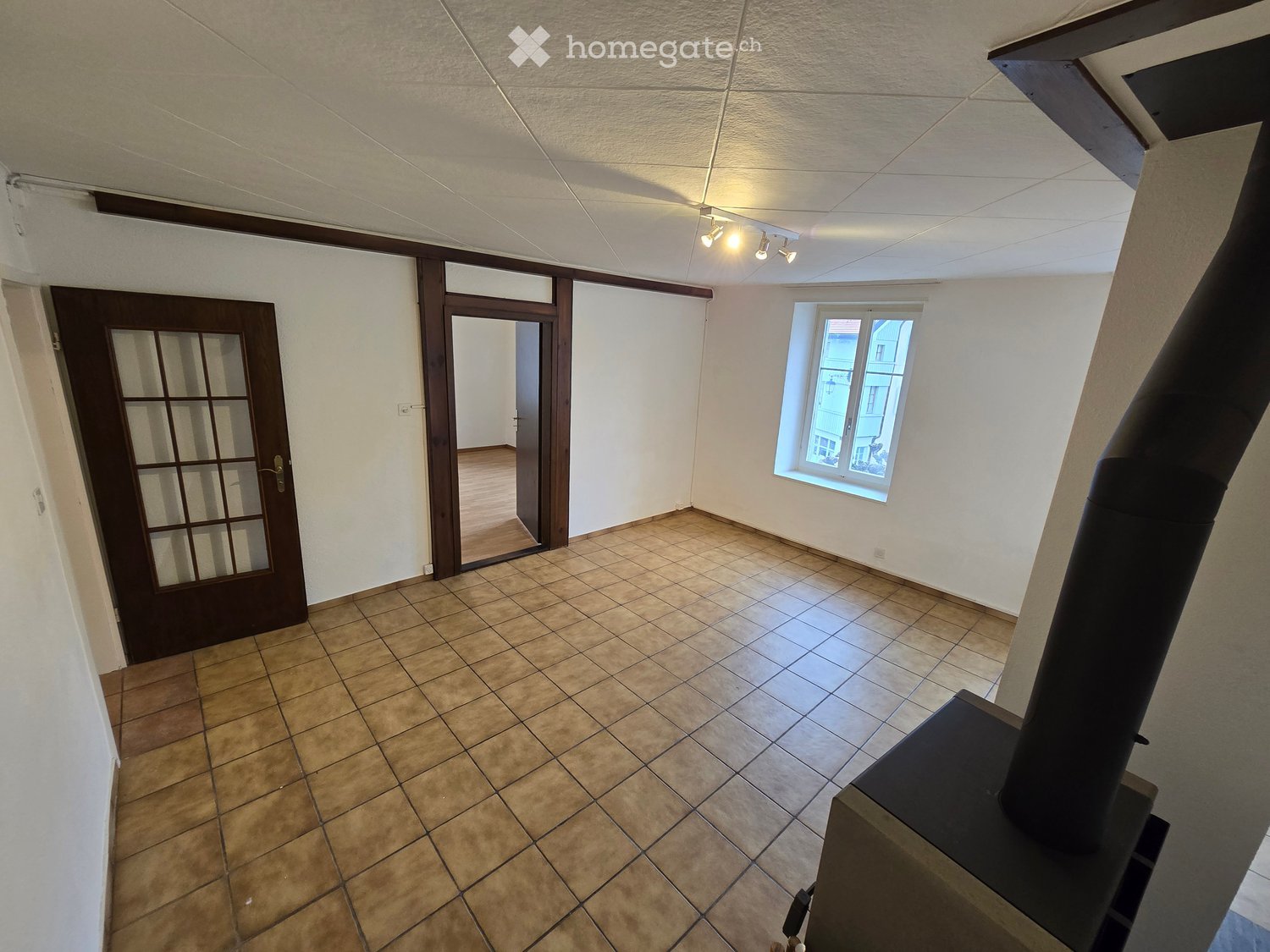Empty room with tiled floor, white walls, a window, and a door leading to another room. A chimney is visible in the foreground.