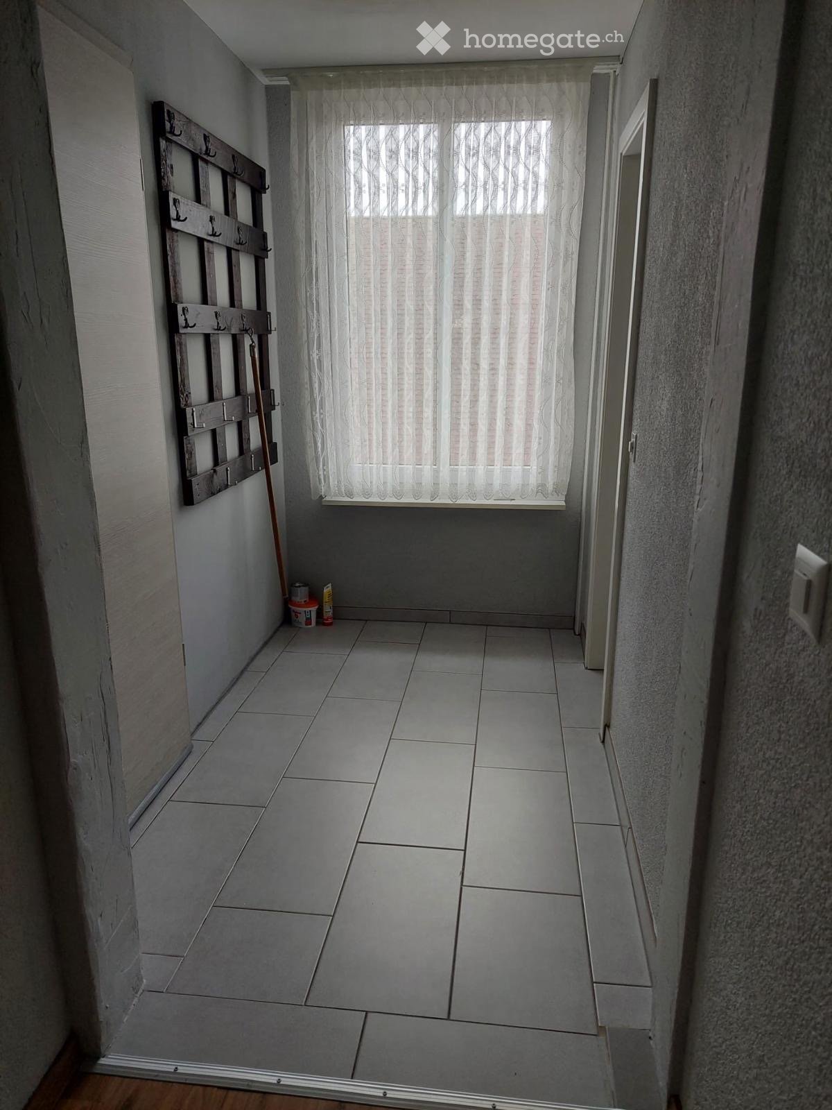 Empty hallway with tiled floor, white walls, and a window with sheer curtains. A coat rack and cleaning supplies are visible on the left side.