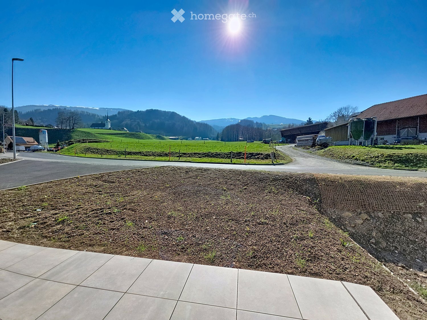 A landscaped outdoor area with a paved walkway, grassy slope, and distant rural buildings and mountains under a clear blue sky.