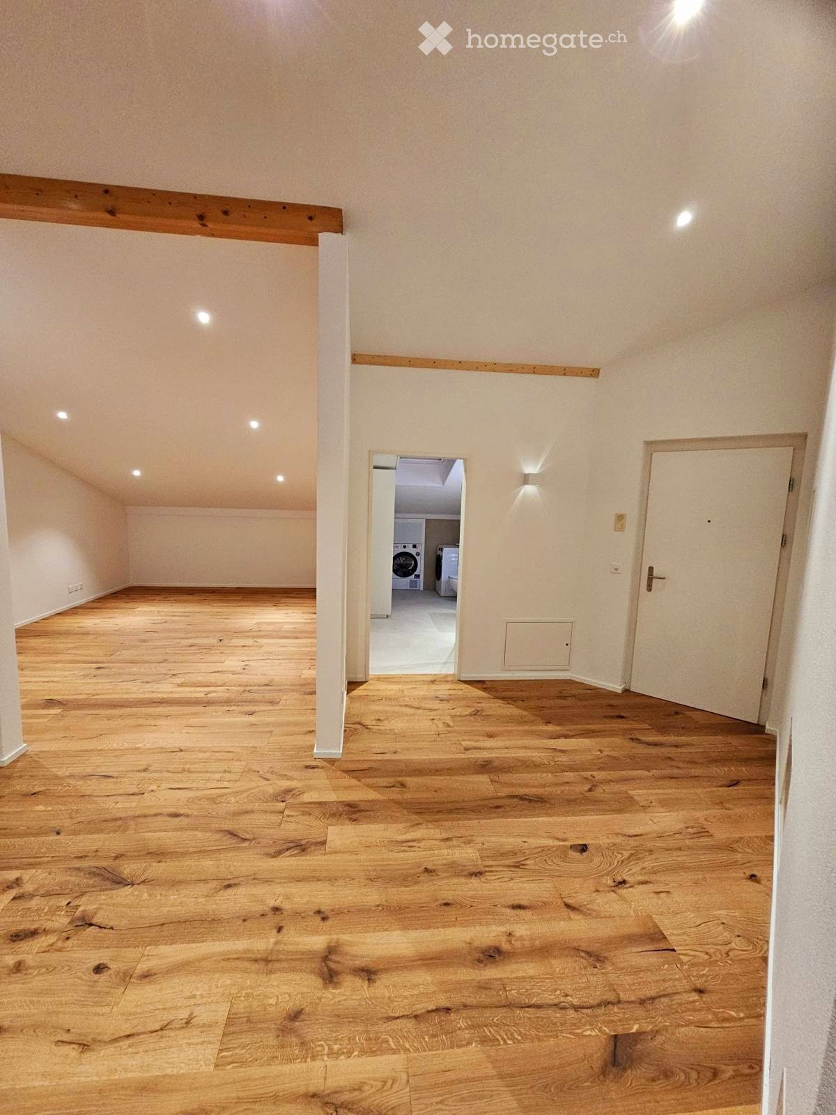 Empty room with wooden flooring, white walls, and recessed lighting. A doorway leads to a small laundry area with a washing machine and dryer.