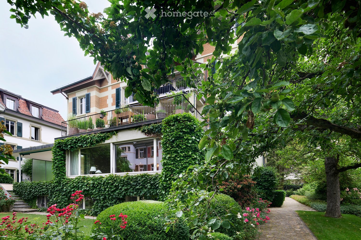 A lush garden area with well-maintained hedges, flowering bushes, and a winding brick pathway. The garden is surrounded by residential buildings with ivy-covered walls and balconies.