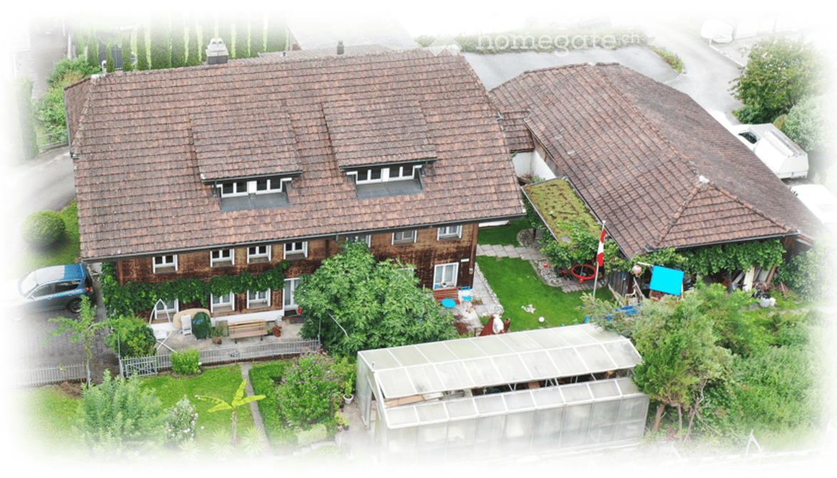 Aerial view of a two-story house with a tiled roof, surrounded by a well-maintained garden, a greenhouse, and a parked car.