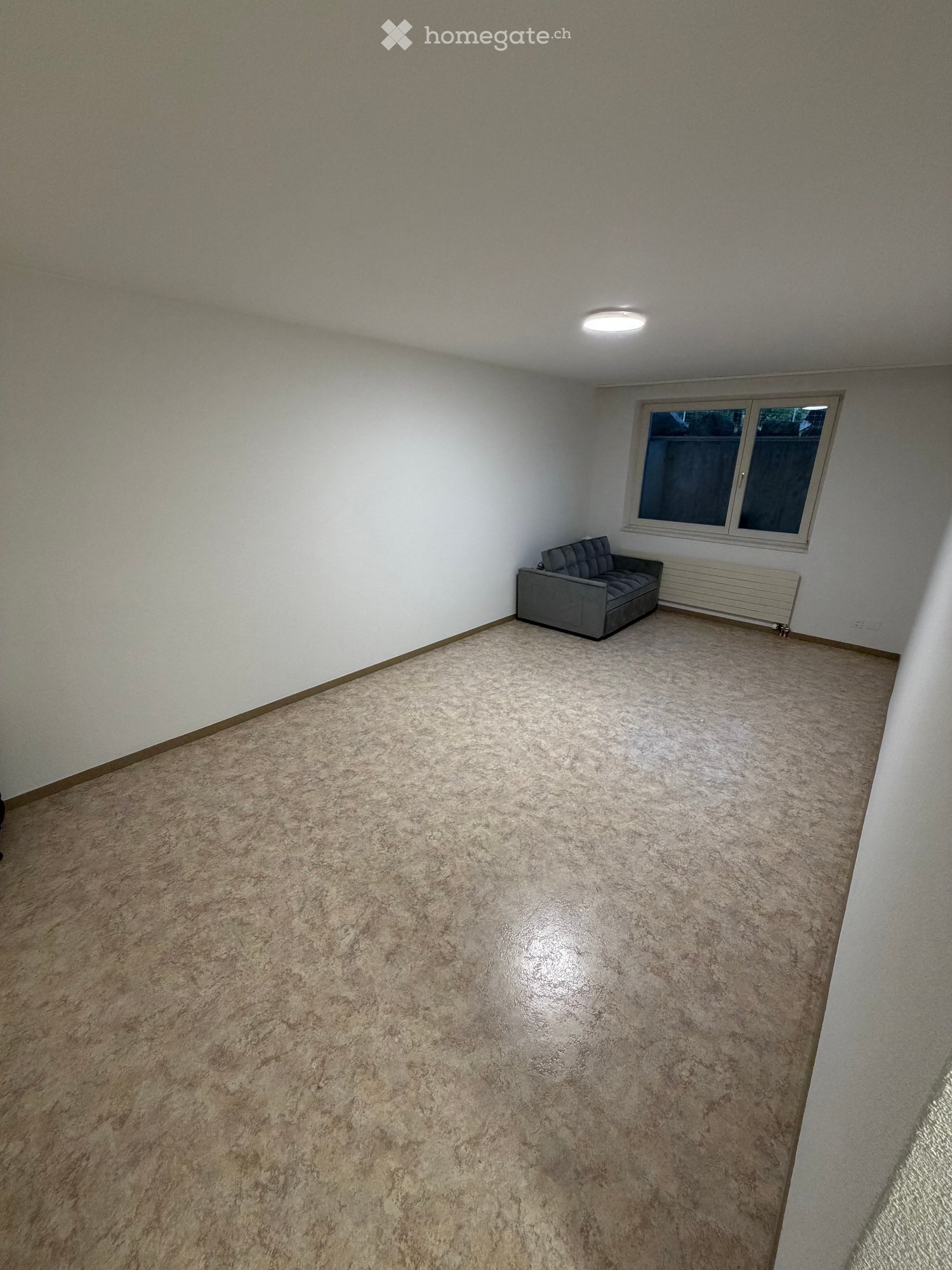 A small, carpeted room with white walls and a single window. Contains a grey sofa against the wall.
