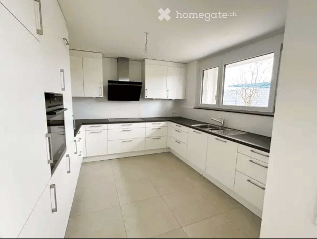 Modern kitchen with white cabinets, black countertops, and stainless steel appliances including an oven and range hood. Large windows provide natural light.
