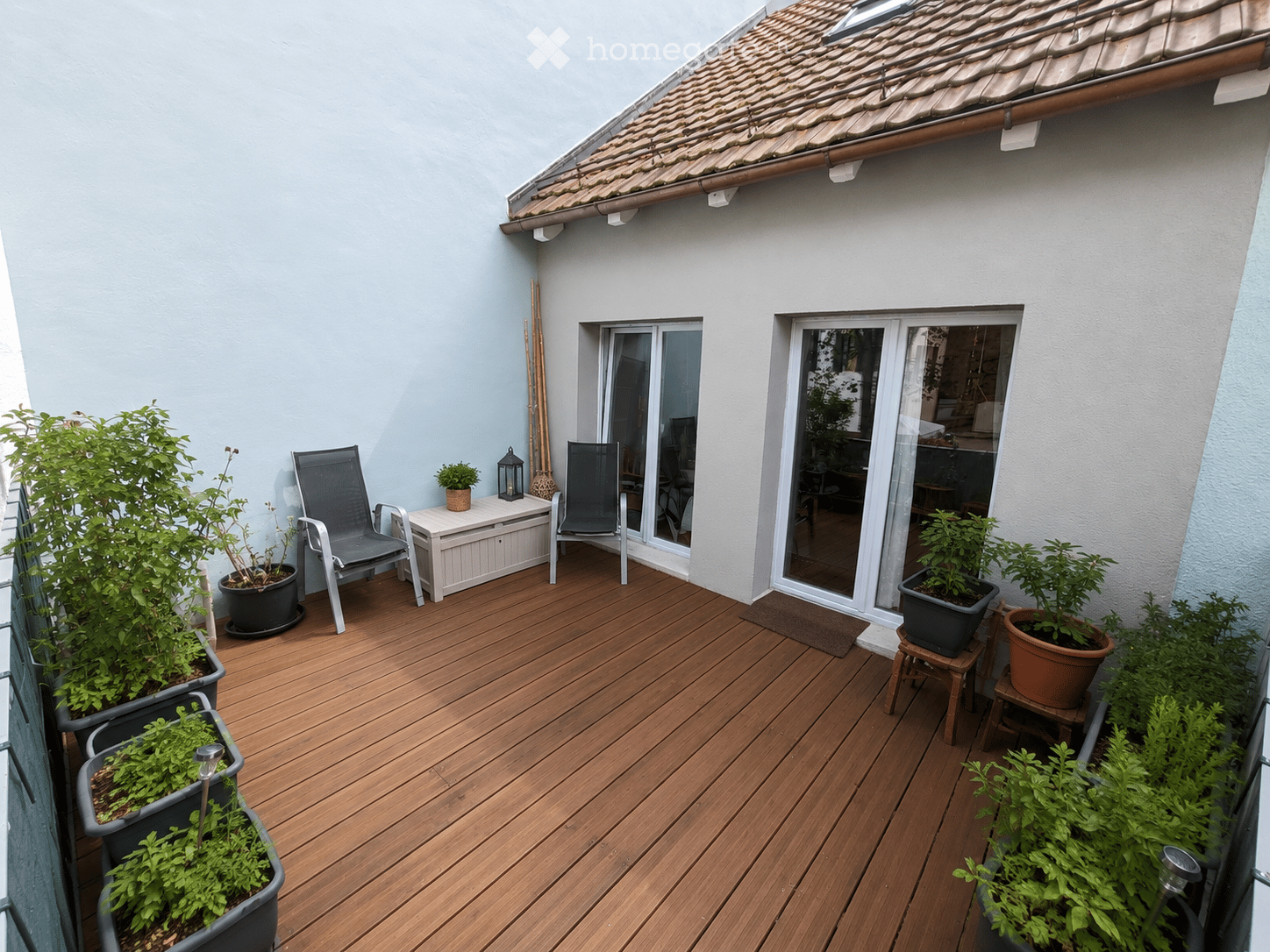 Wooden deck terrace with two chairs, a small table, potted plants, and a lantern. The terrace has a slanted roof and overlooks a building entrance.