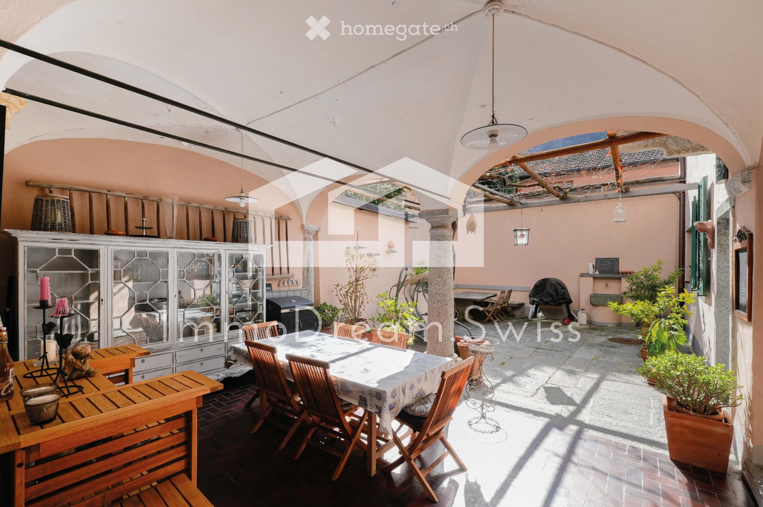 Courtyard with wooden furniture, dining table, chairs, potted plants, and a covered seating area with hanging lights.