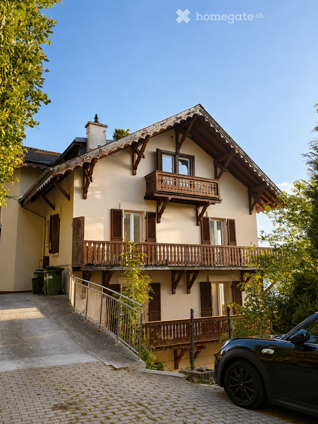 Two-story house with beige walls, wooden balconies, and multiple windows with shutters. A black car is parked on the driveway.