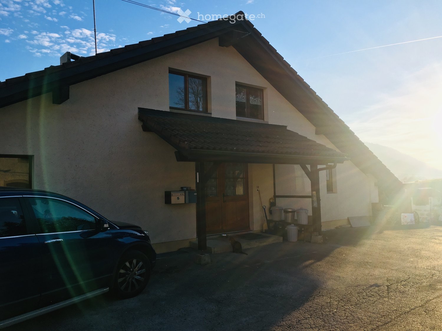 Two-story house with a tiled roof, white walls, and a covered entrance area. A car is parked in front of the house.