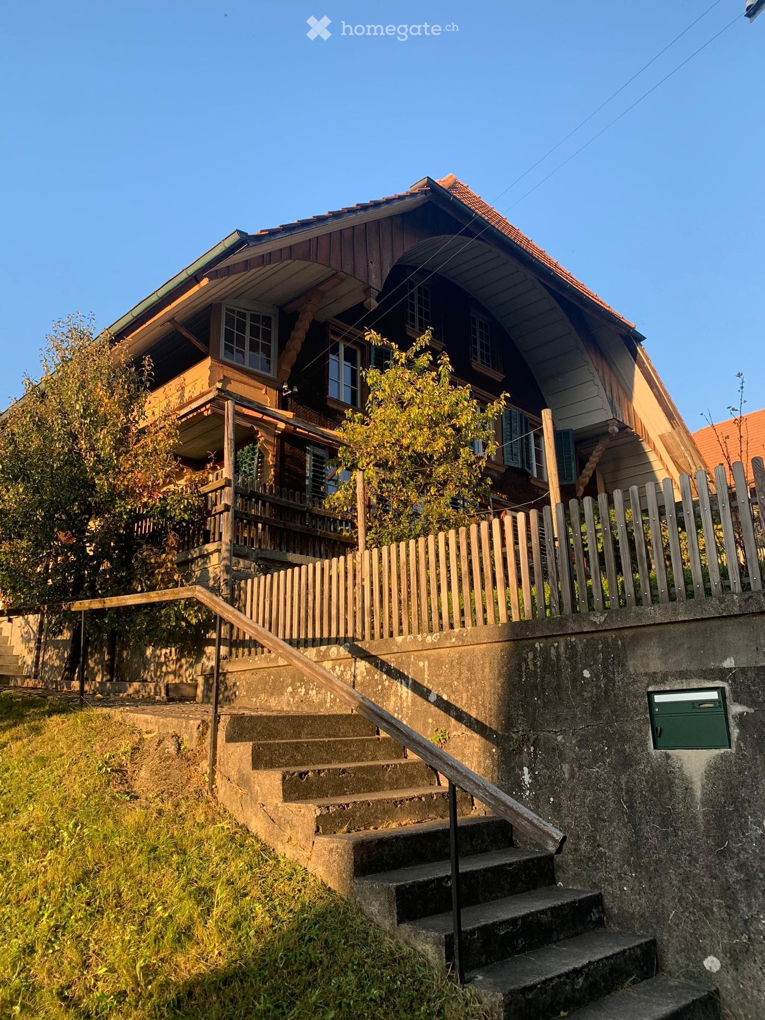 Two-story wooden house with a sloped roof, multiple windows, and a wooden balcony, surrounded by a wooden fence and greenery.