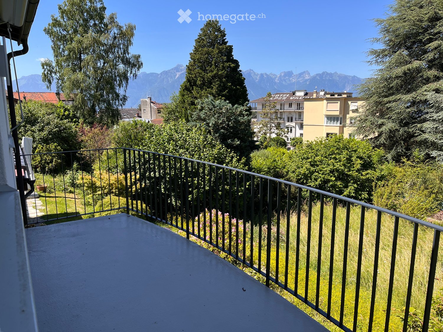 Balcony with metal railing overlooking a green area with trees and distant mountains.