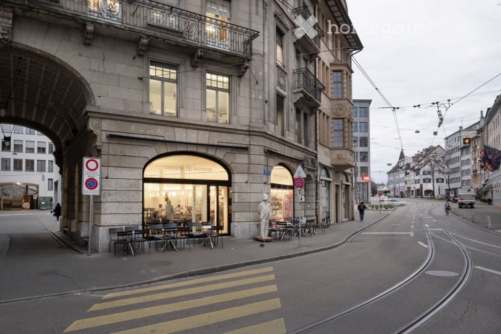 Urban street view with a multi-story building featuring large windows, balconies, and a storefront with outdoor seating. The street includes traffic signs, a pedestrian crossing, and a tram line.