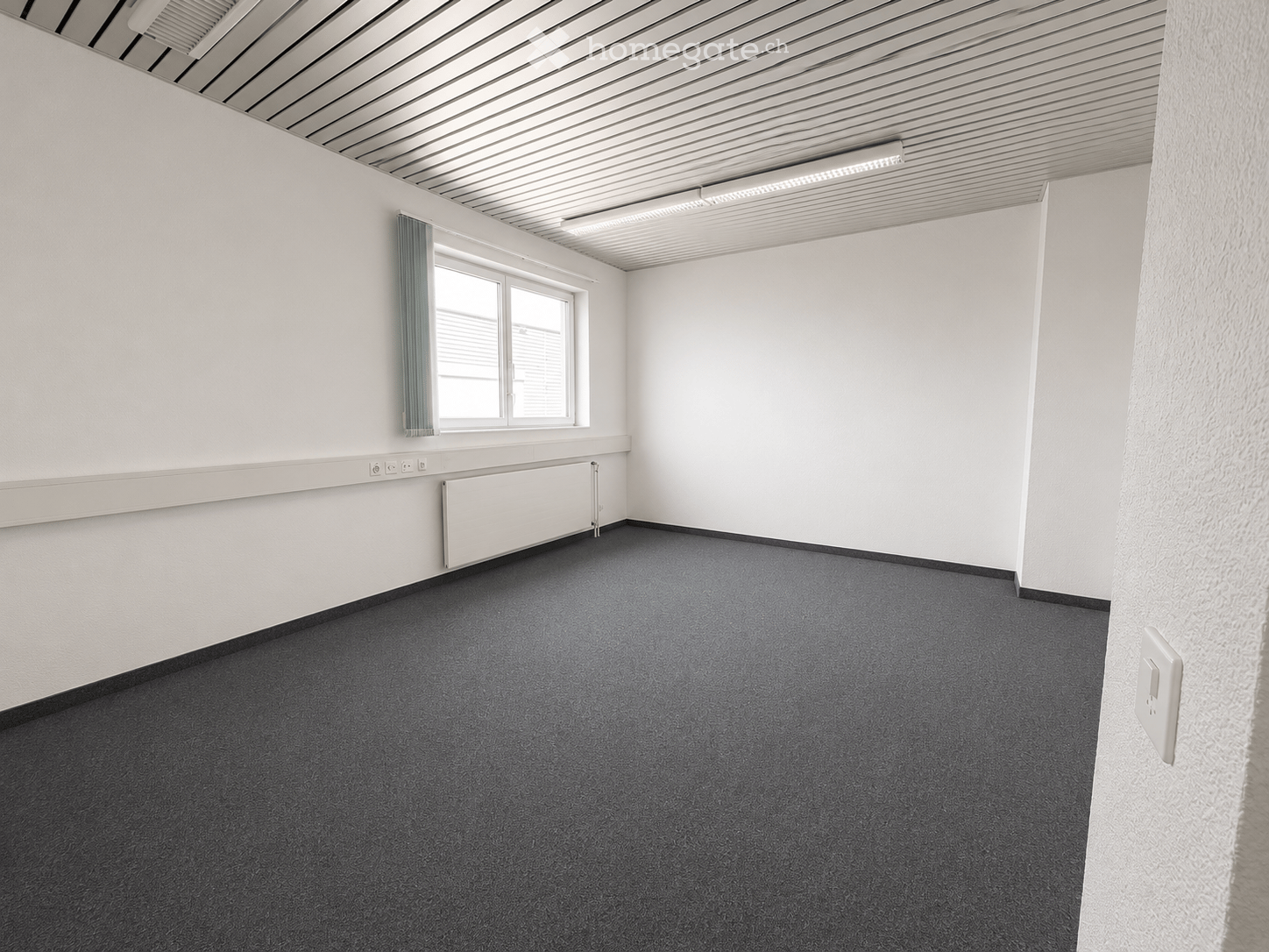 Empty room with white walls, grey carpet, a window with a green curtain, and a radiator below the window.
