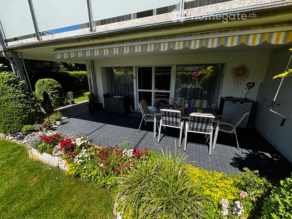 Outdoor patio area with a dining set, striped chairs, and a table, surrounded by a well-maintained garden with colorful flowers and shrubs.