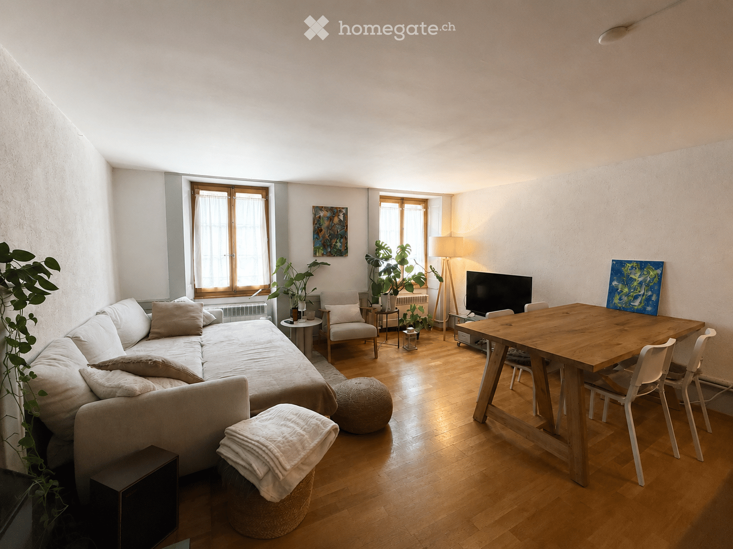 A modern living room with a sofa, coffee table, armchairs, a round wooden table with chairs, a floor lamp, and a television. The room features wooden flooring, large windows, and decorative plants.