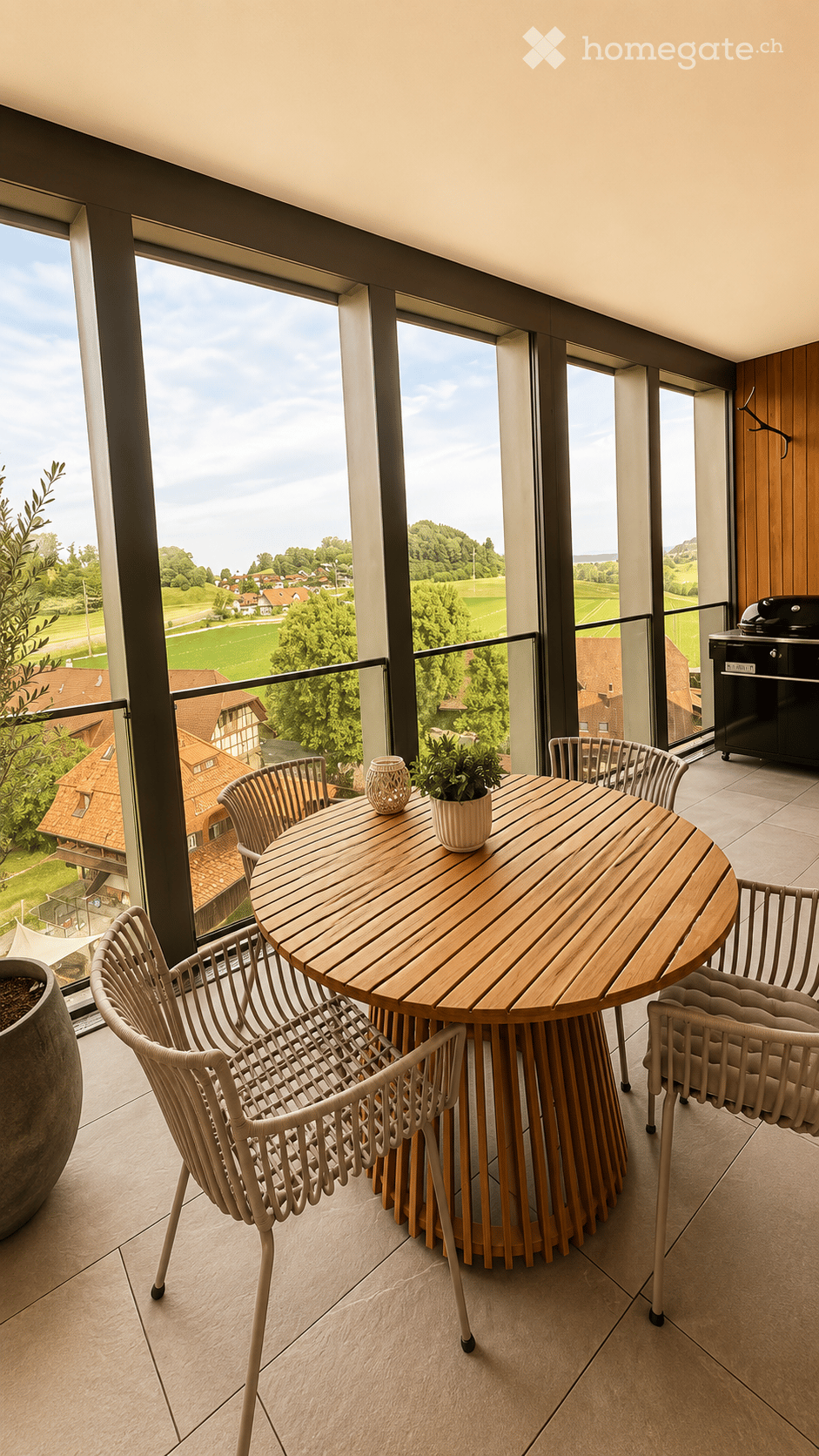 Balcony with a round wooden table, four white wicker chairs, a small potted plant, and a decorative vase. Large windows offer a scenic view of a countryside landscape.