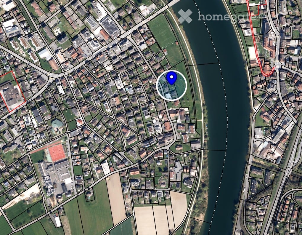 Aerial view of a residential area with streets, buildings, and a river running alongside. A blue marker indicates a specific location.