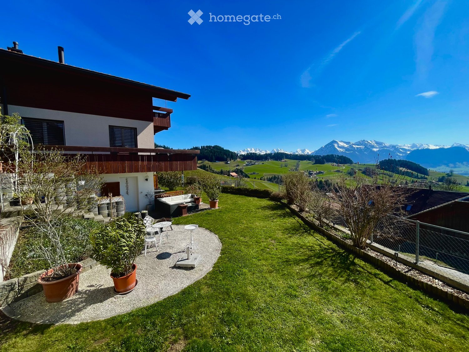 Garden / Outdoor Area. Garden with a well-maintained lawn, patio seating area with a sofa, table, and chairs, surrounded by potted plants and a scenic mountain view in the background.