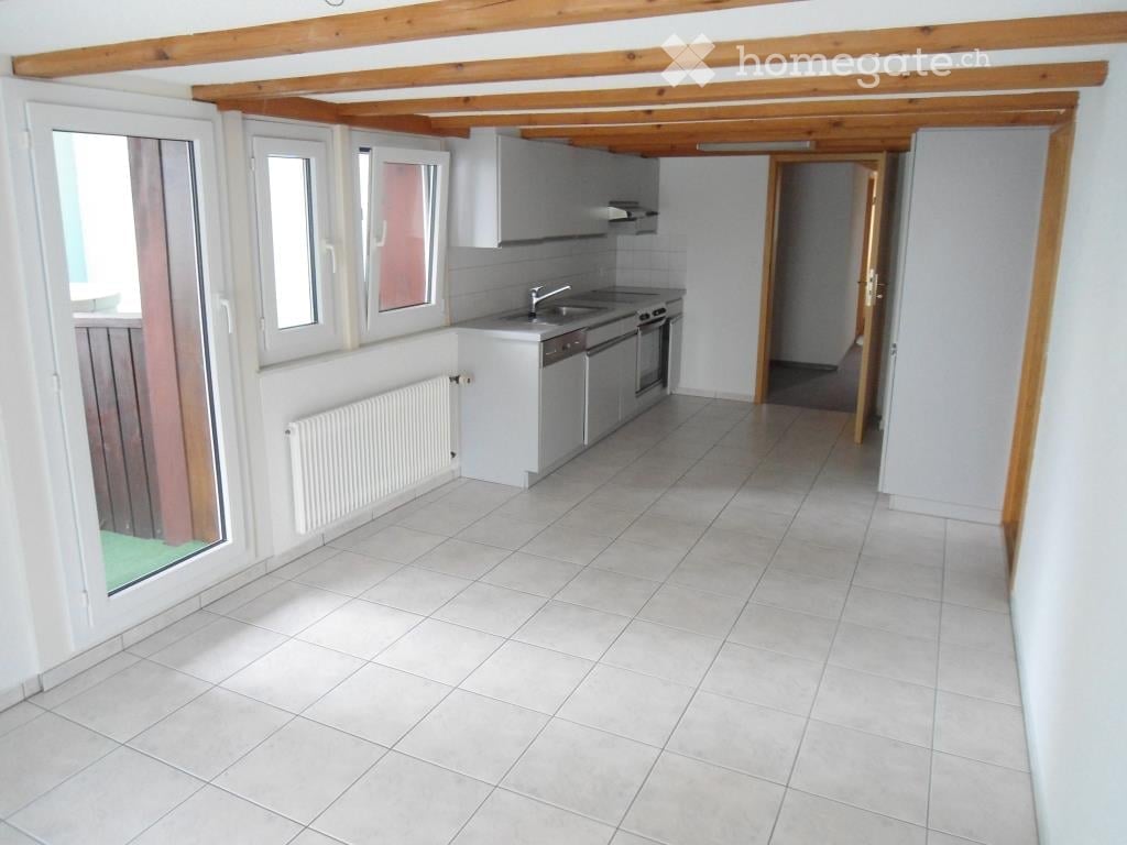Bright, modern kitchen with white tiled flooring, white cabinets, and stainless steel appliances including a sink, faucet, oven, and dishwasher. A radiator is visible on the wall, and the space has large windows allowing natural light.