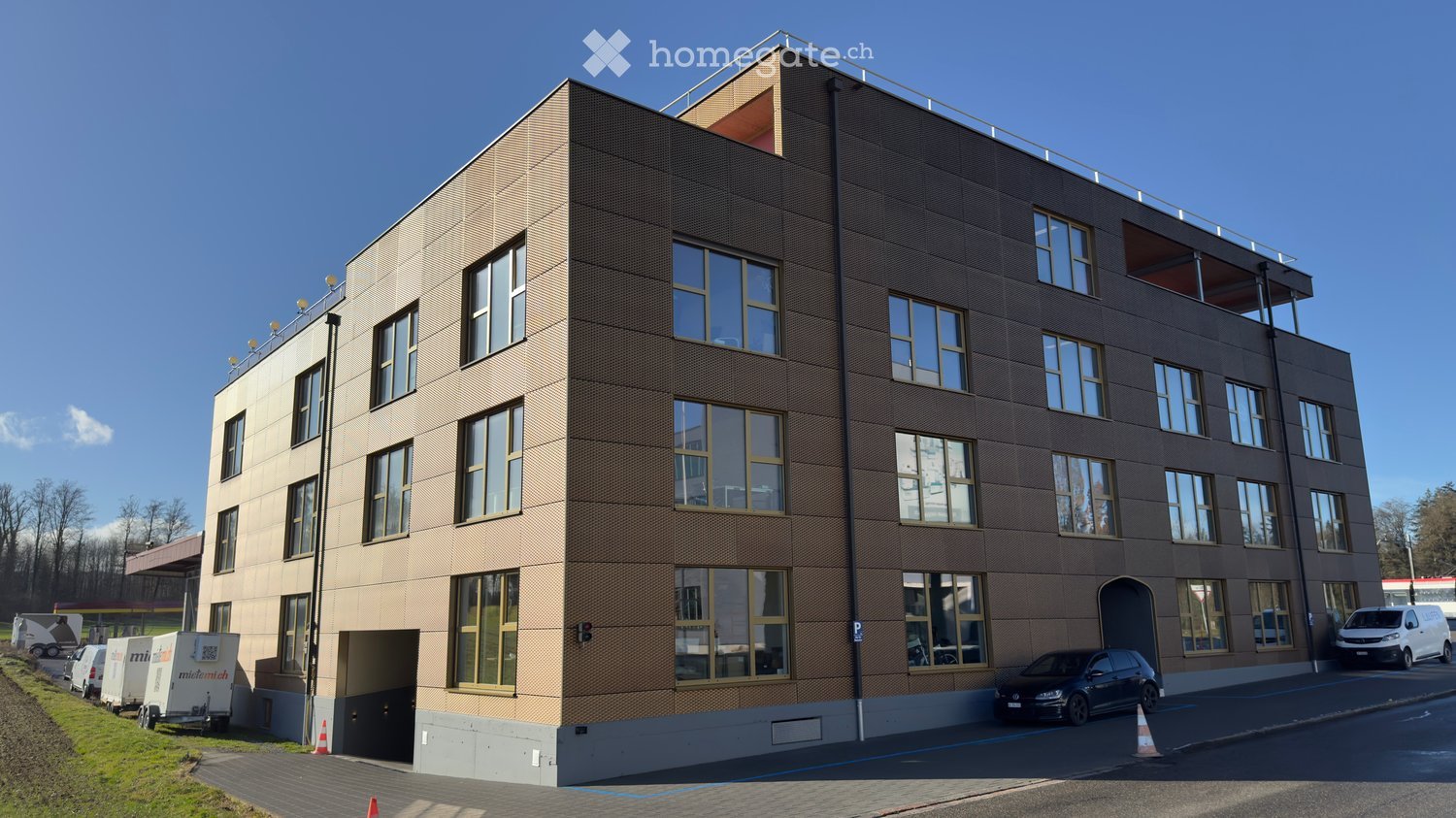 Modern multi-story building with a textured facade, large windows, and a rooftop terrace. Several cars are parked in front, and there is a trailer and a van nearby.