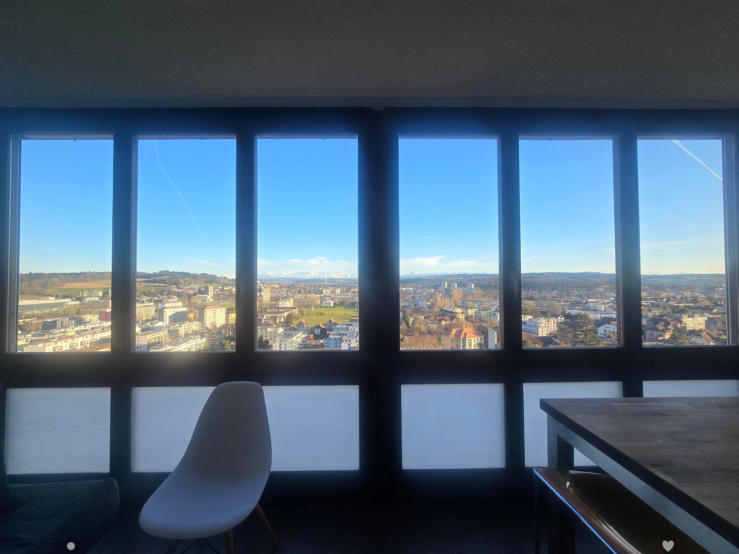A modern, minimalist room with large windows offering a panoramic city view. The room features a sleek white chair and a wooden table.