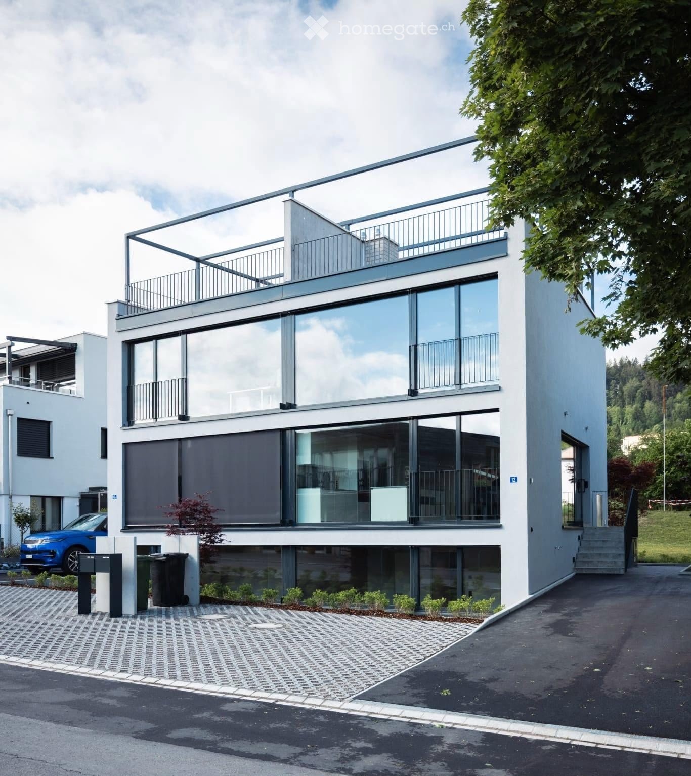 Modern two-story house with large glass windows, balconies, and a raised ground floor. Features a paved driveway, small garden area, and a blue car parked nearby.
