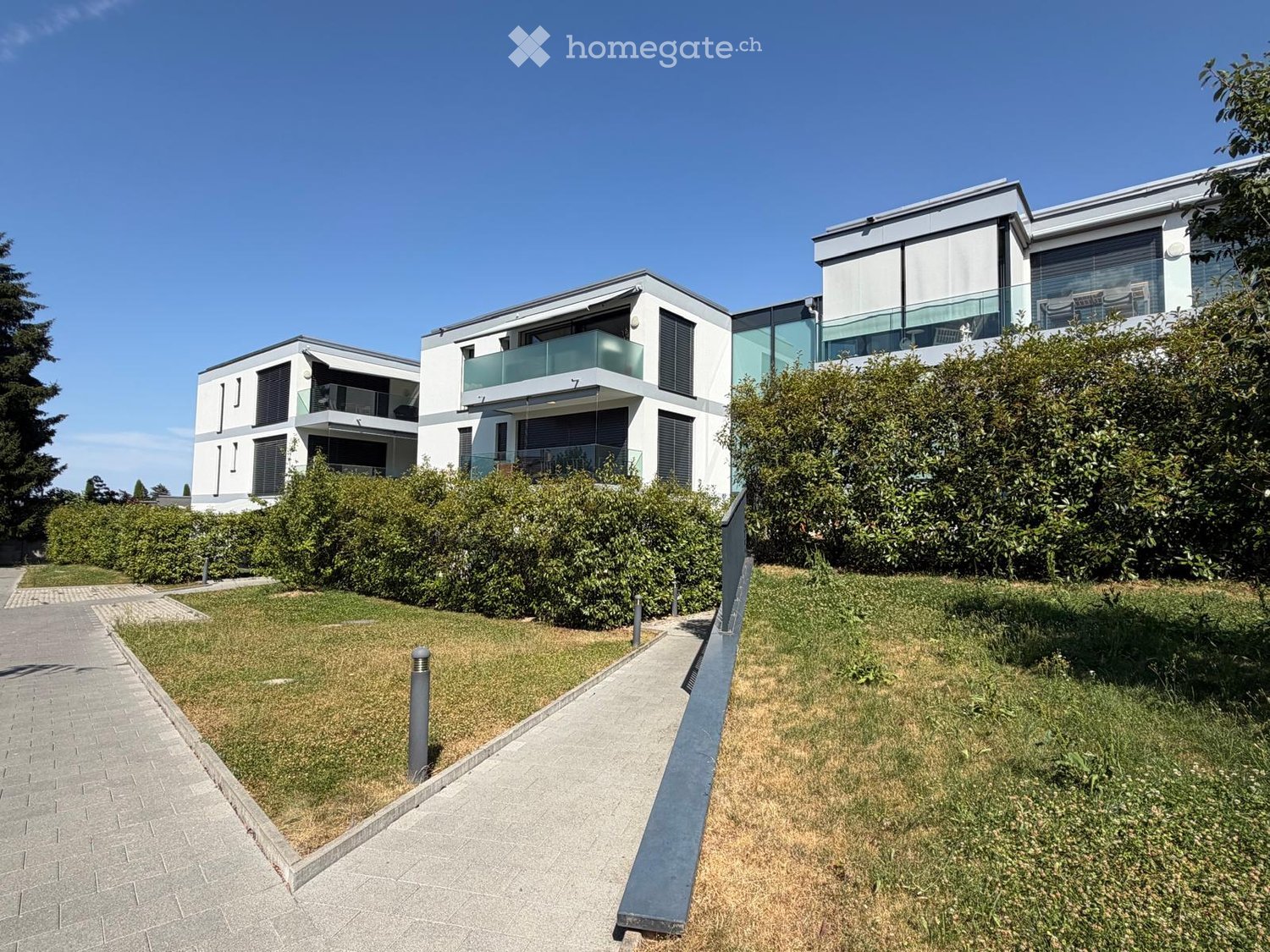 Modern two-story residential building with white and grey facade, large windows, and balconies surrounded by greenery and a paved pathway.