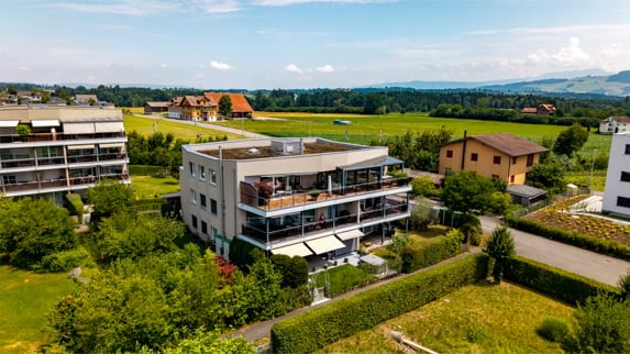 Modern multi-story residential building with balconies and large windows, surrounded by greenery and other houses in a rural setting.