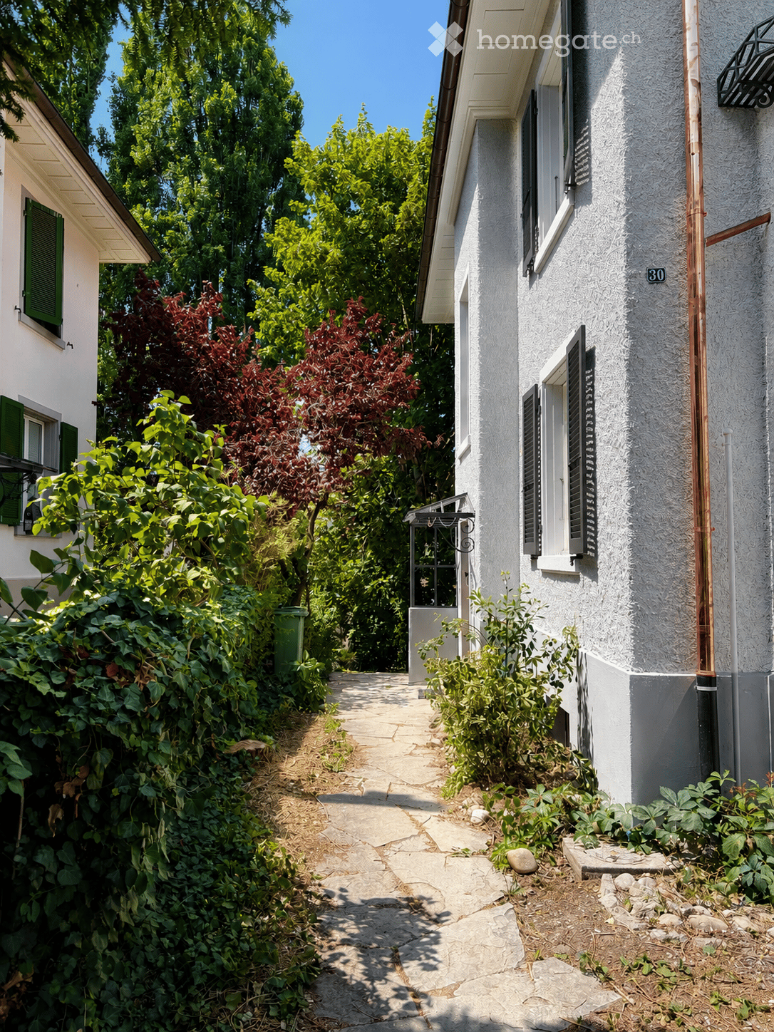A narrow garden pathway lined with greenery leads to a white house with green shutters and a small balcony. The house has a textured facade and a visible address number30.