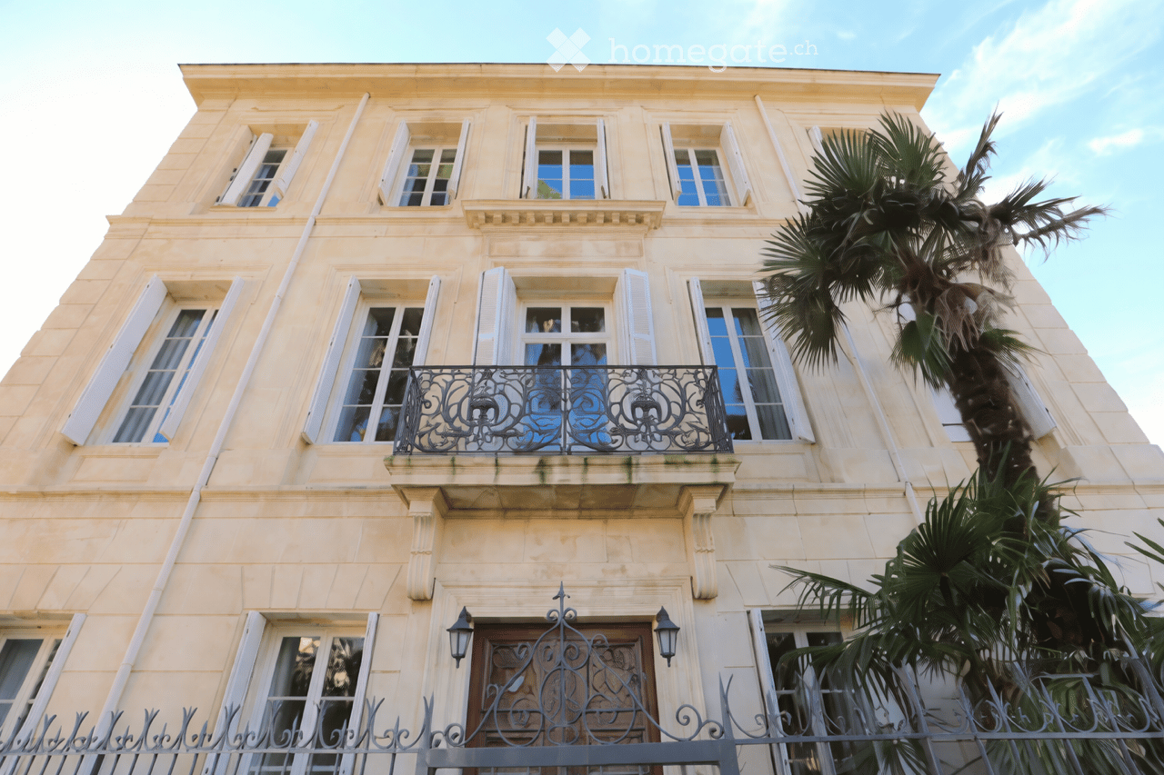 A multi-story building with light-colored stone facade, multiple windows with shutters, ornate wrought-iron balcony, and palm trees in front.
