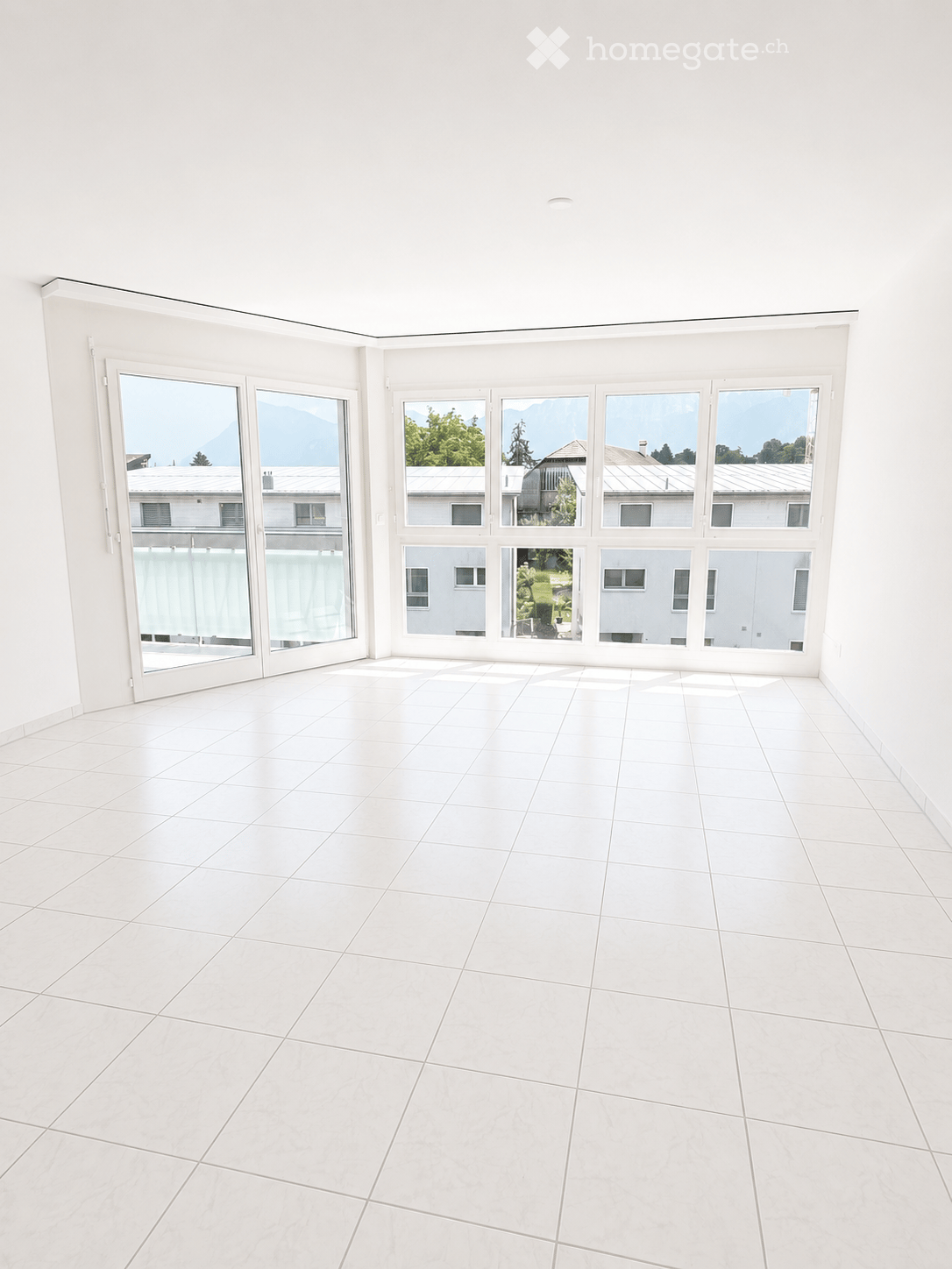 Empty room with white tiled floor and large windows offering a view of the surrounding area and mountains.