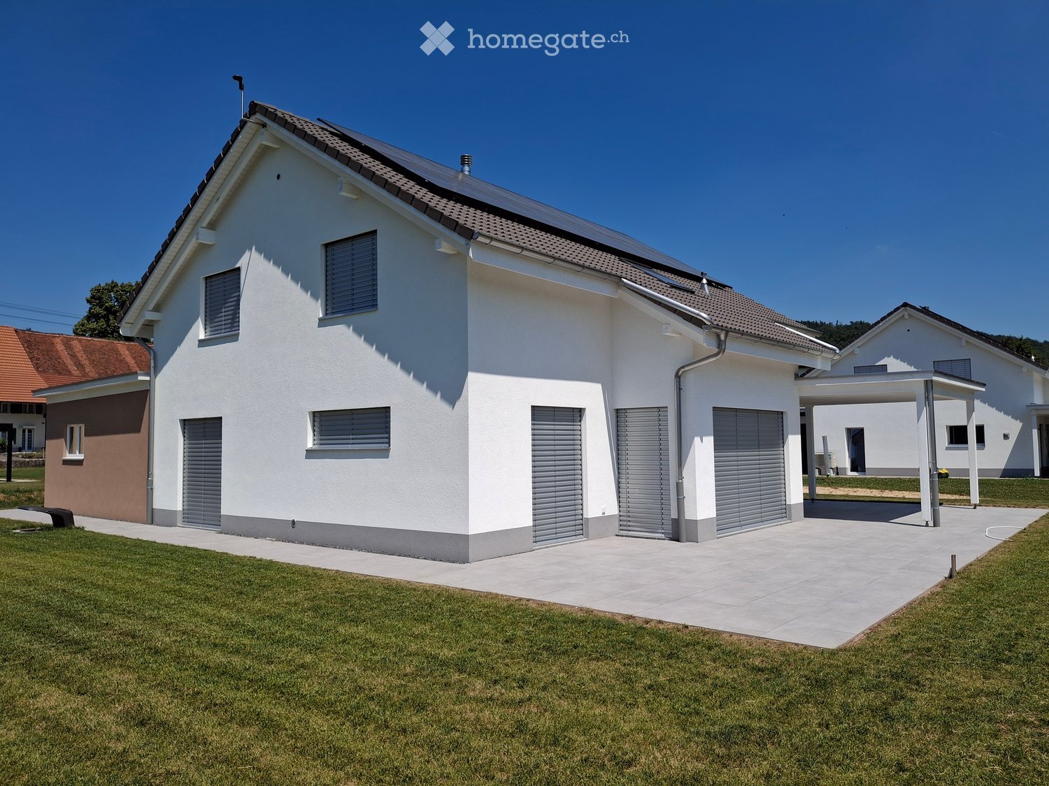 Modern two-story house with white exterior, grey shutters, and a covered driveway. The house features a garage and is surrounded by a well-maintained lawn.