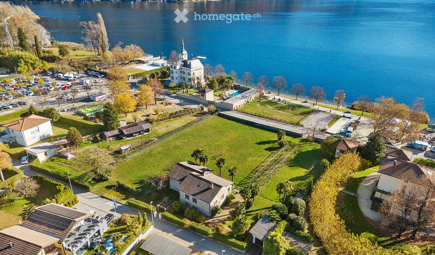 Aerial view of a lakeside area with a large grassy field, several buildings, parking lot, and a view of the lake.