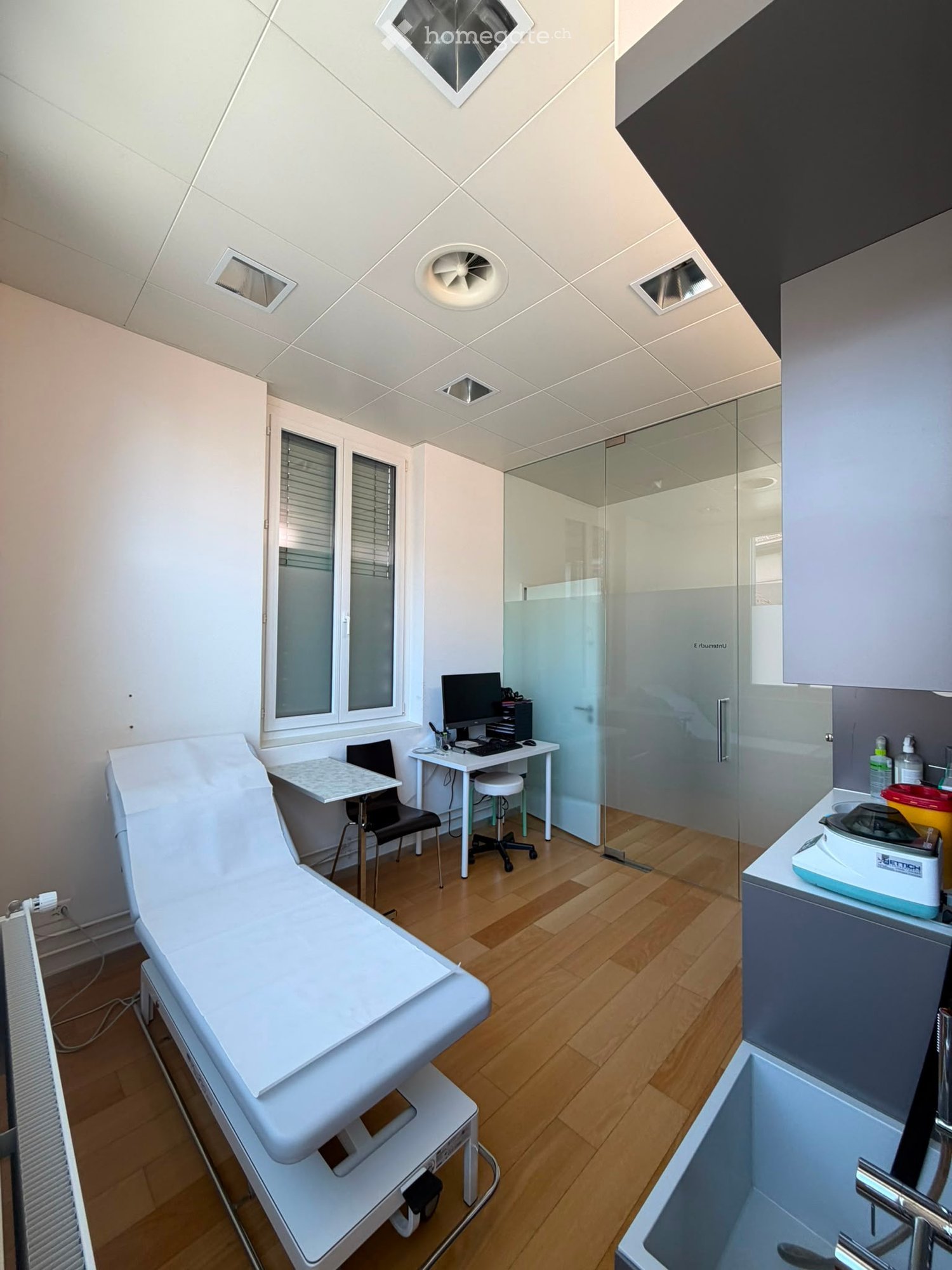 Modern medical examination room with a white exam table, desk with computer, chair, sink, and cabinets.