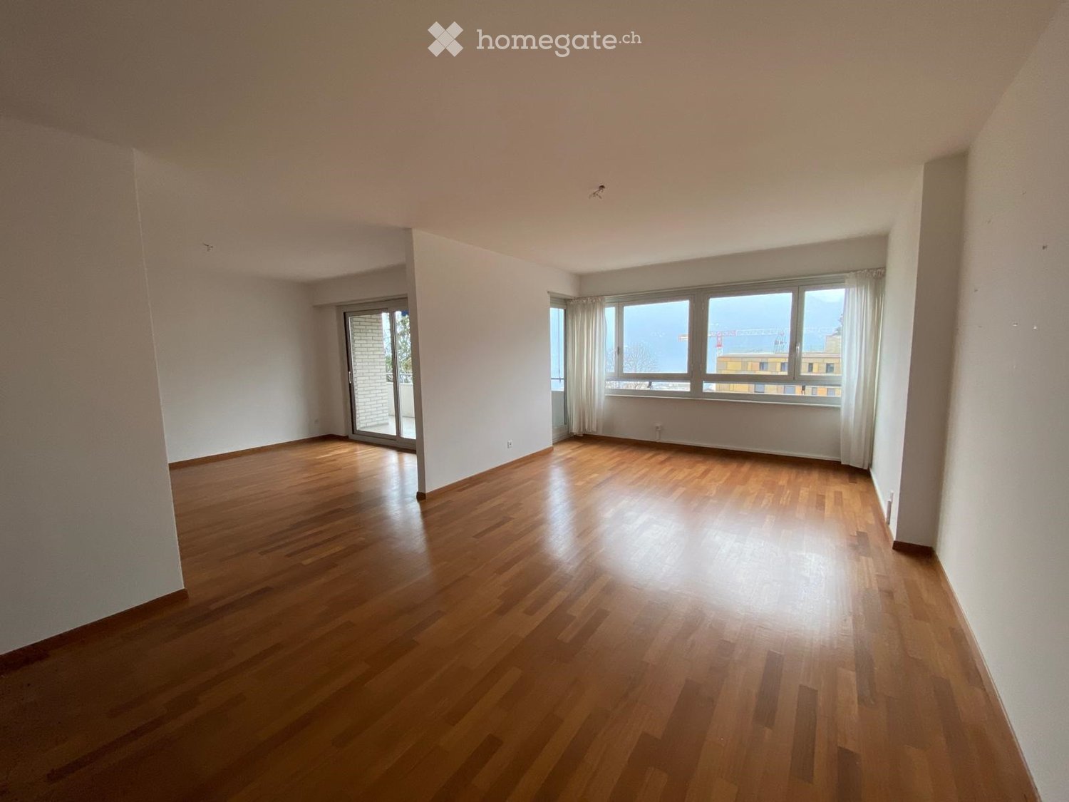 Empty room with wooden flooring, white walls, large windows with curtains, and a sliding glass door leading to a balcony.