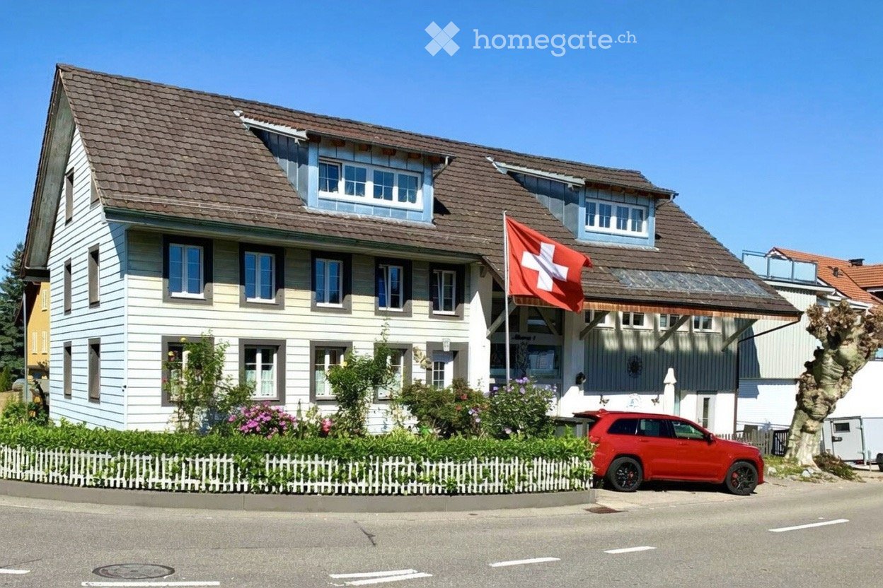 Two-story house with a brown tiled roof, white siding, multiple windows, and a Swiss flag. A red SUV is parked in front, and there is a small garden with a white picket fence.