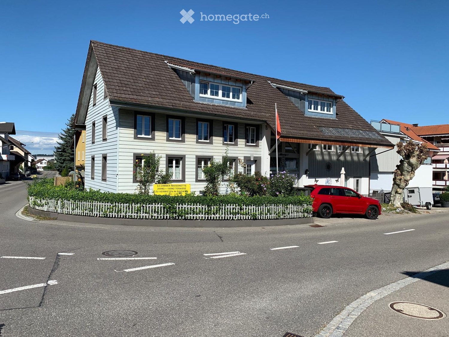 Two-story house with a brown tiled roof, white siding, multiple windows, and a small garden with a white picket fence. A red SUV is parked on the street in front of the house.