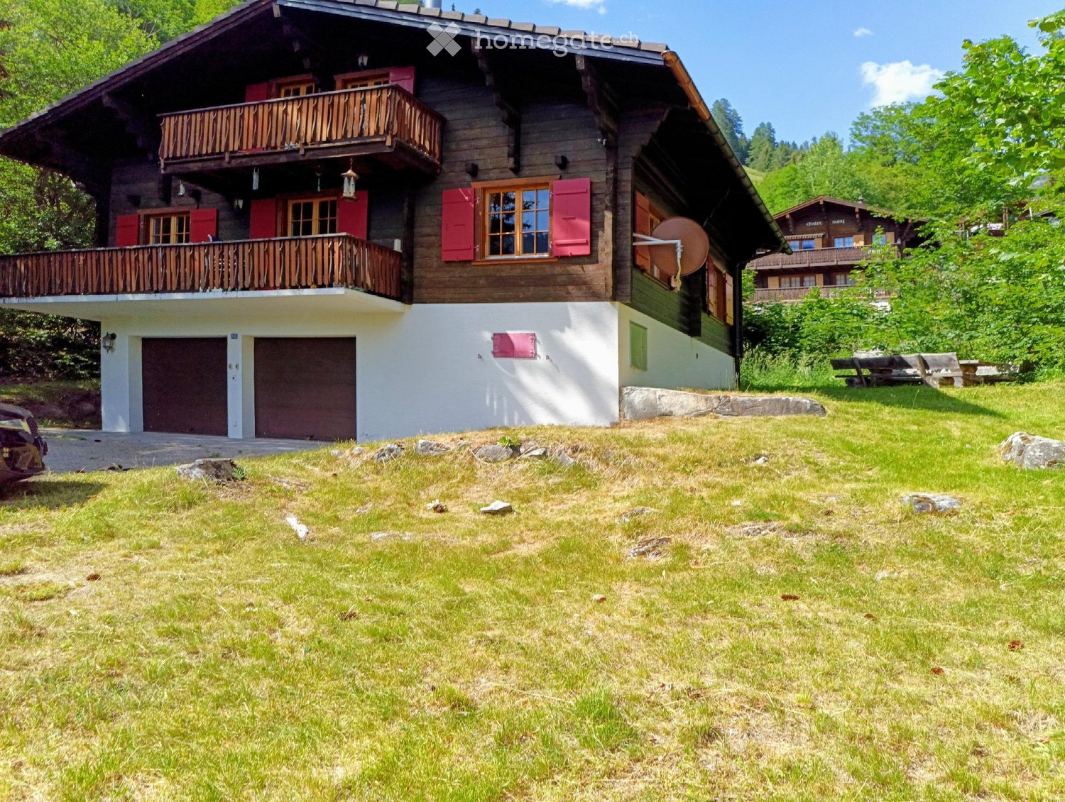 Two-story wooden house with red shutters, balconies, and a garage, surrounded by green grass and trees.