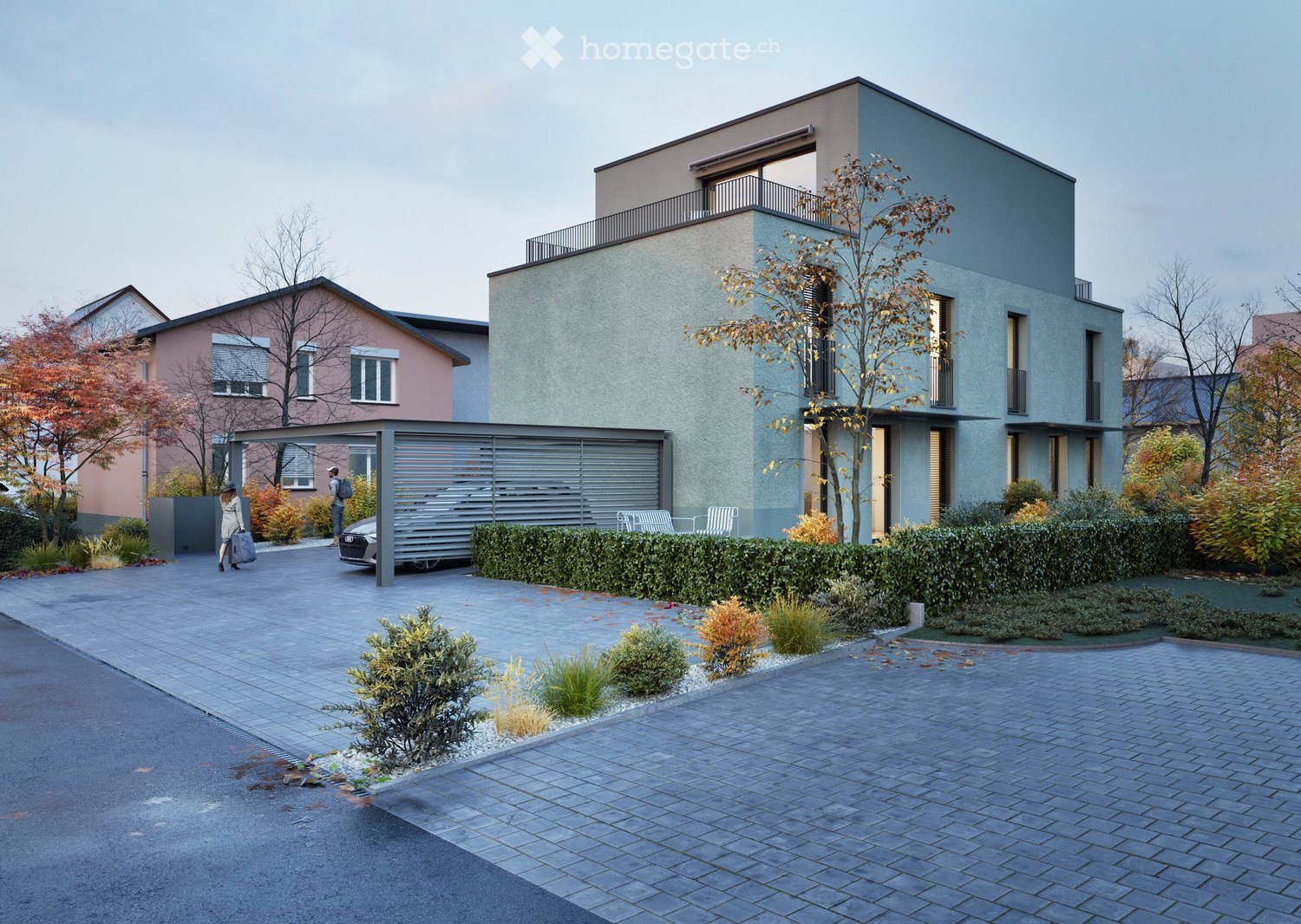 Modern two-story house with grey facade, large windows, and a balcony on the upper floor. The house is surrounded by a well-maintained garden with shrubs and trees, and a paved driveway leads to a covered parking area.