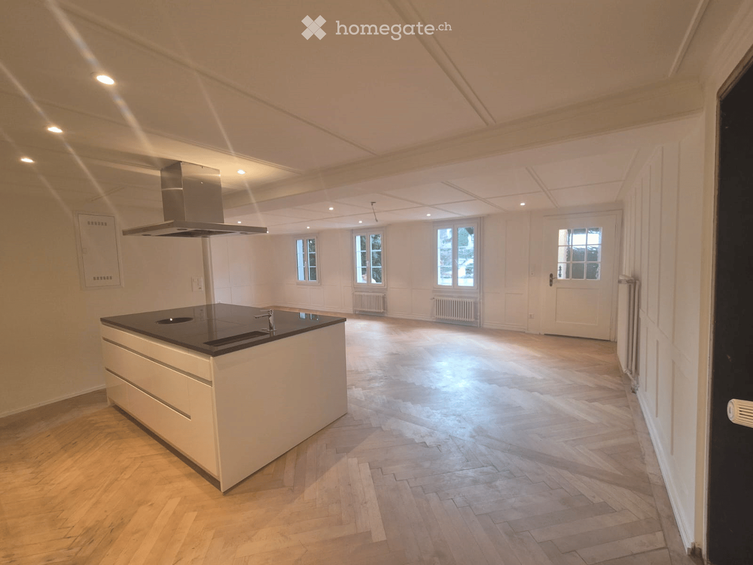 Open kitchen with island, wooden parquet floor, white walls, ceiling with recessed lighting, windows, kitchen hood, central island with sink and stove