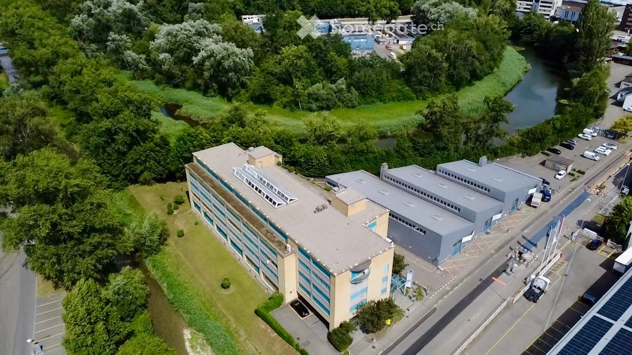 Aerial view of a large industrial or commercial building complex with multiple sections, surrounded by greenery and a river, with parking areas and vehicles visible.