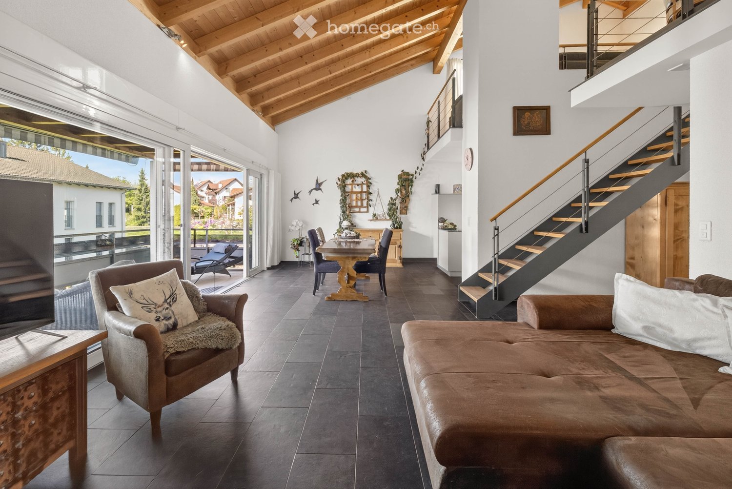 Modern living room with exposed wooden ceiling beams, large windows, and a staircase leading to an upper level. Features include a brown leather sofa, a wooden armchair with a deer pillow, a wooden dining table with chairs, and decorative wall art.