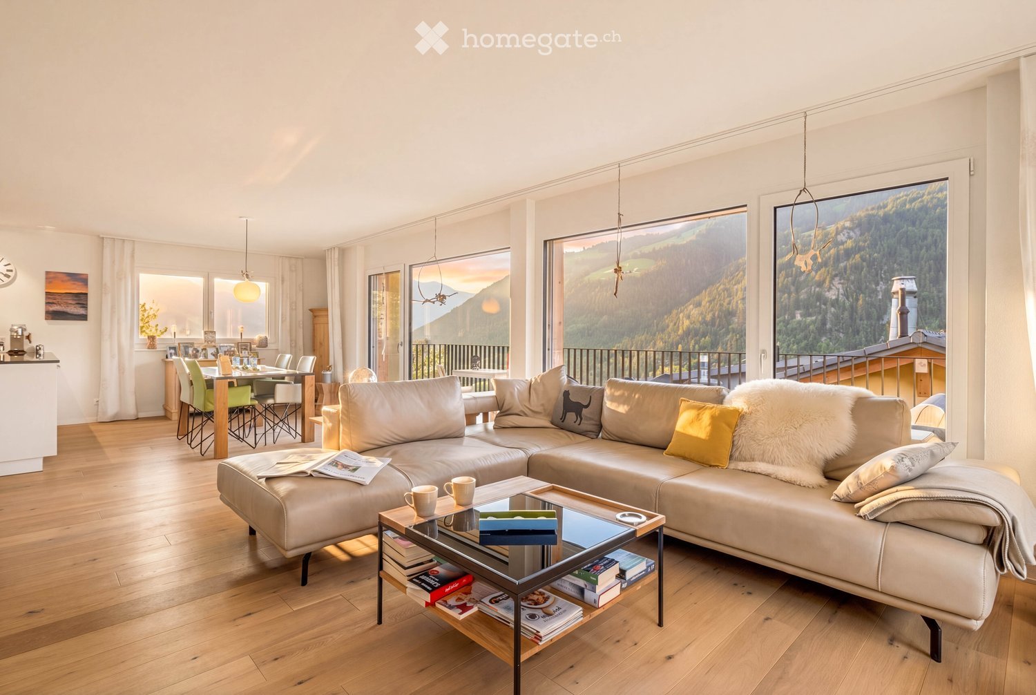 Bright living room with large windows offering a scenic mountain view, featuring a cream-colored sectional sofa, a glass coffee table with books and cups, a dining area with chairs and a table, and modern decor.