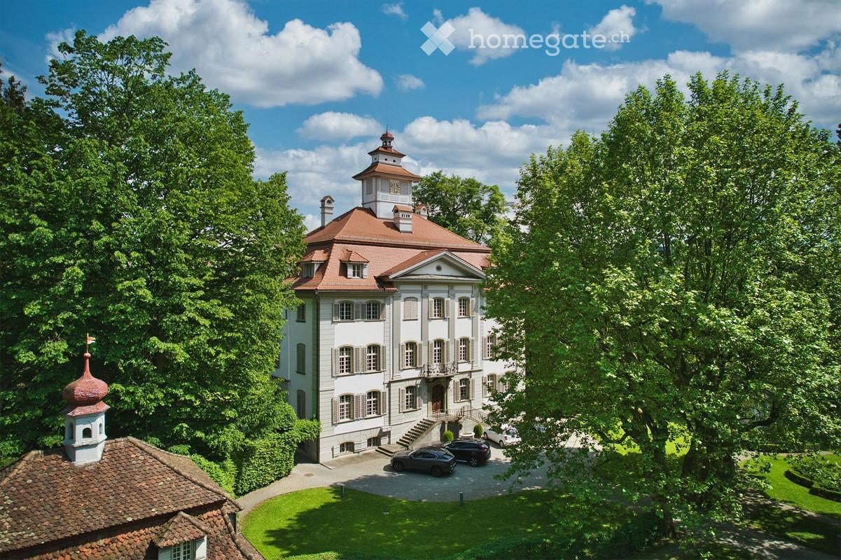 A large, elegant white mansion with a red-tiled roof, surrounded by lush greenery and trees. The mansion features multiple windows, a central entrance with steps, and a small tower with a dome on the roof. Several cars are parked in the driveway.