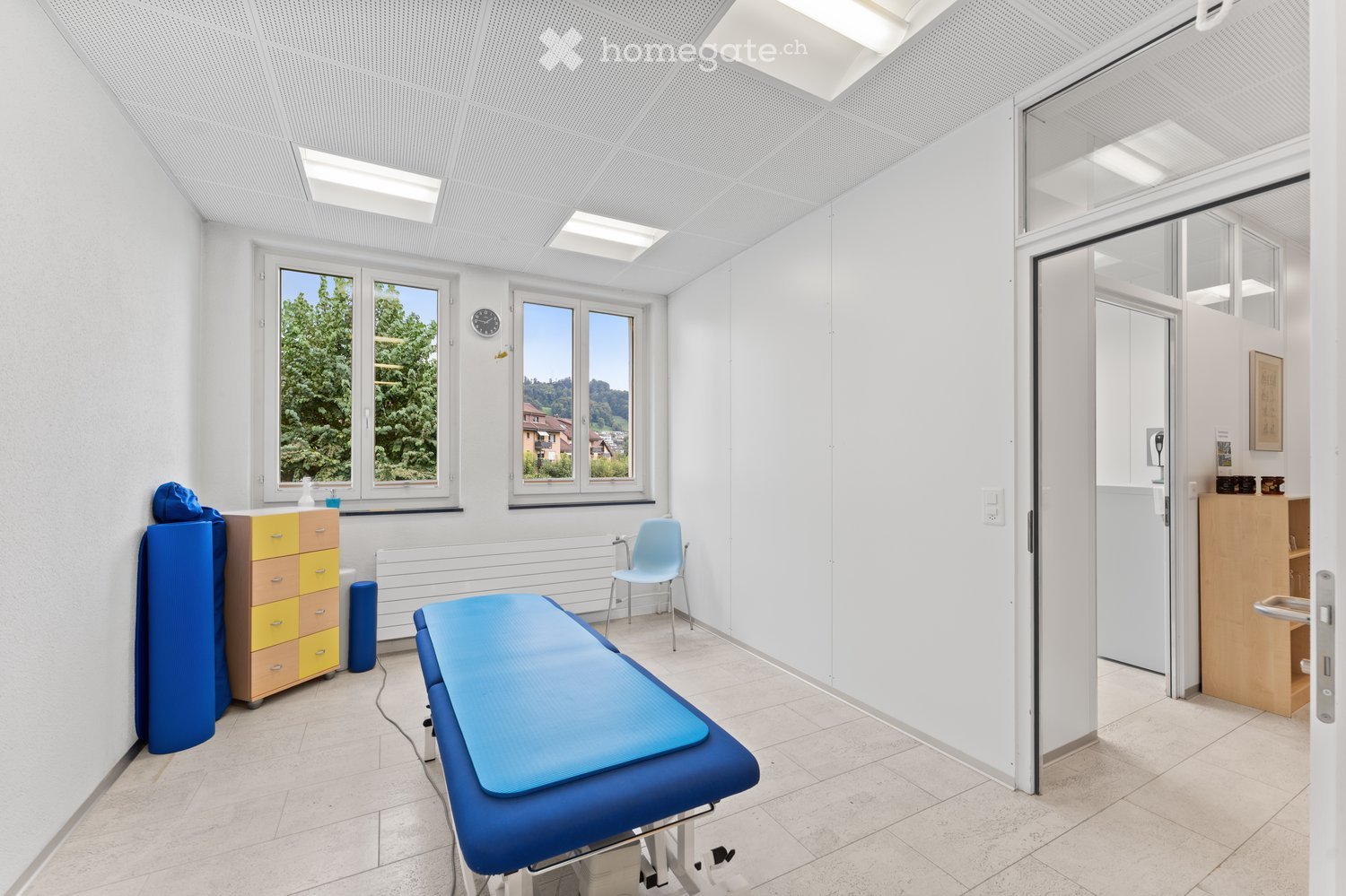 A clean, well-lit examination room with a blue examination table, a light blue chair, a yellow and brown cabinet, and a clock on the wall. Large windows offer a view of greenery outside.