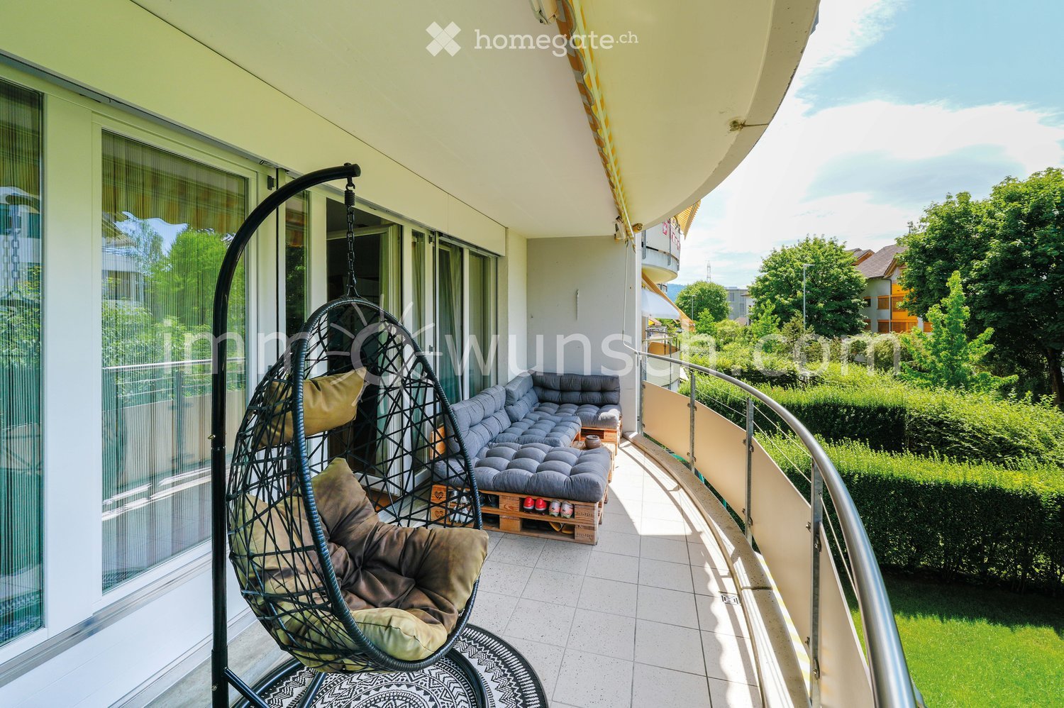 Balcony with a modern design, featuring a grey sectional sofa, a wicker hanging egg chair with cushions, a small wooden table, and a patterned rug.